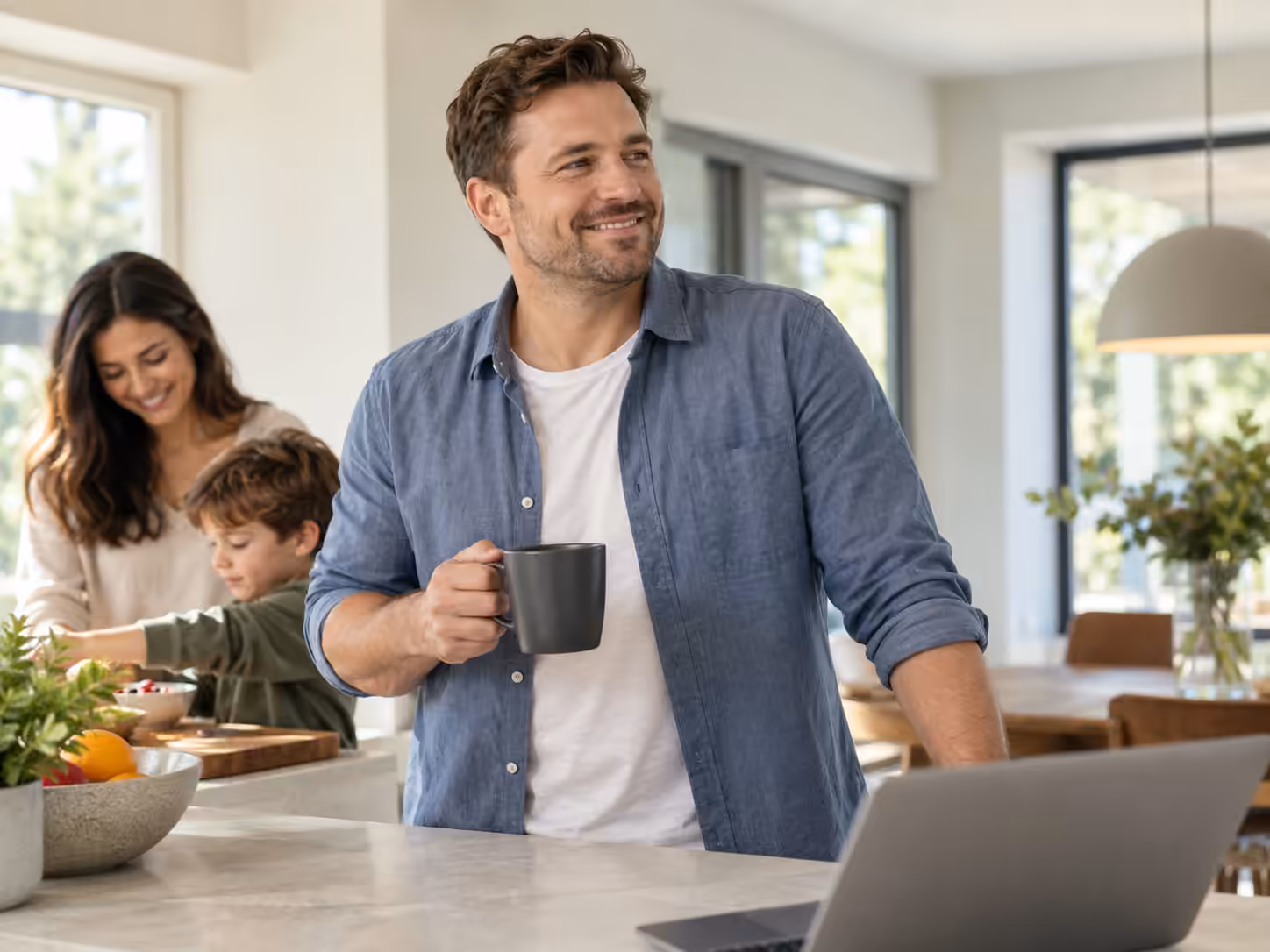 An adult man holding a mug in a bright family kitchen, with his partner and child in the background.